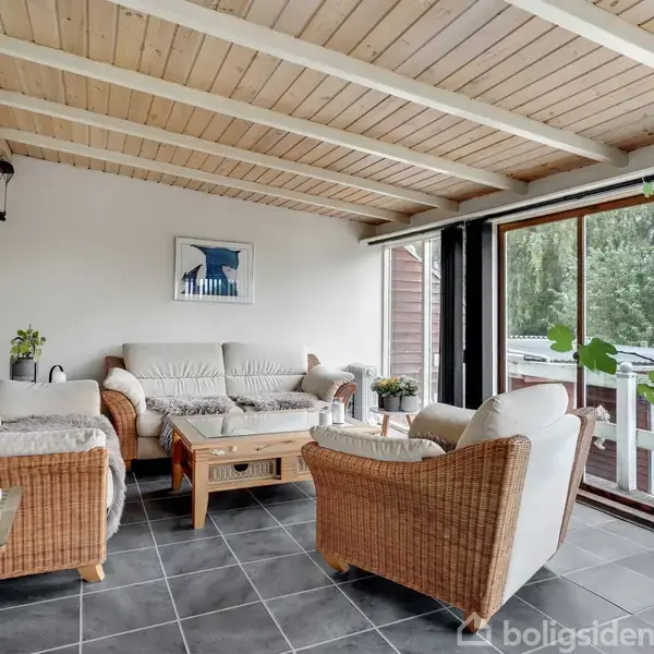 Fletmøbler omkranser et sofabord i en lys havestue. Store vinduer åbner op til udsigt over en terrasse med træer. Loftet er af træ, og gulvet flisebelagt.