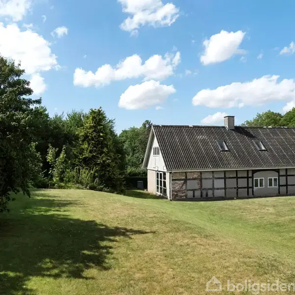 Et bindingsværkshus står stille på en stor græsplæne, omgivet af træer og buske under en blå himmel med spredte hvide skyer.