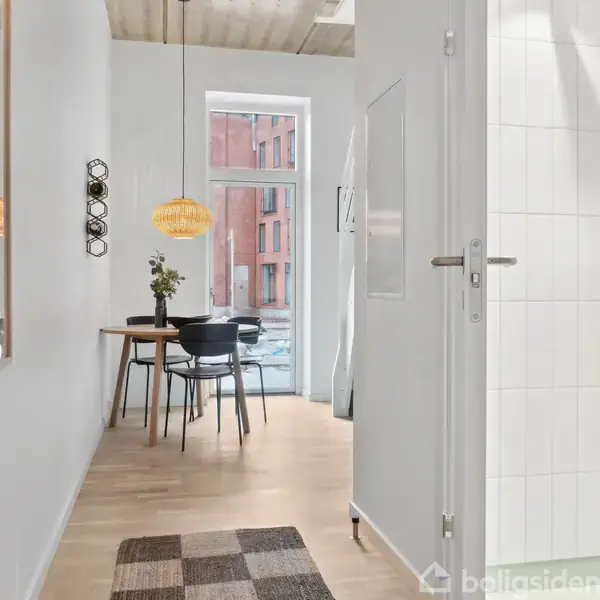 En gang fører til et lyst køkken med træbord og sorte stole. En loftslampe hænger over bordet. Vinduet viser en udsigt med røde bygninger. En dør åbner til badeværelse.