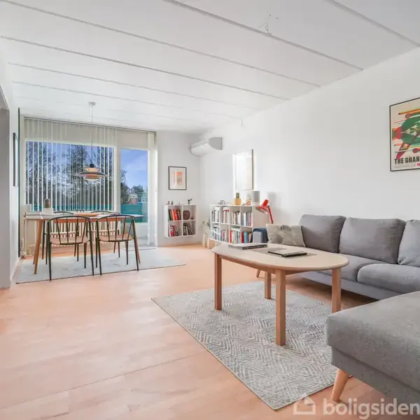 Stue med grå sofa og sofabord, står stillet ved gråt tæppe. Spisebord med seks stole ses i baggrunden nær vinduer med persienner. Væggene er pyntet med billeder.