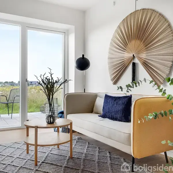 Sofa med farverige puder står i en lys stue. Rundt sofabord med vase, moderne kunst på væggen. Store vinduer med udsigt til terrasse og grønne marker.