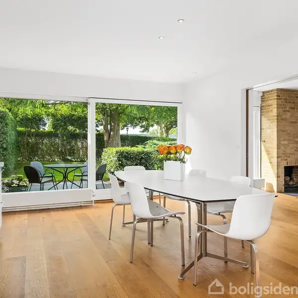 Et lyst køkken med hvidt spisebord og stole står foran store vinduer, der viser en have. Der er friske blomster på bordet, og en anden stue ses gennem en åbning.