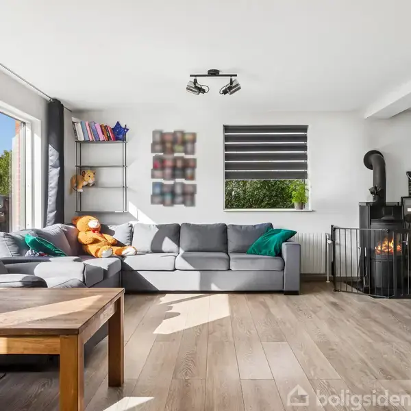 Grå sofa i en stue, omgivet af farverige puder og bamser. Solskin skinner gennem store vinduer til venstre. En brændeovn til højre skaber en hyggelig atmosfære.