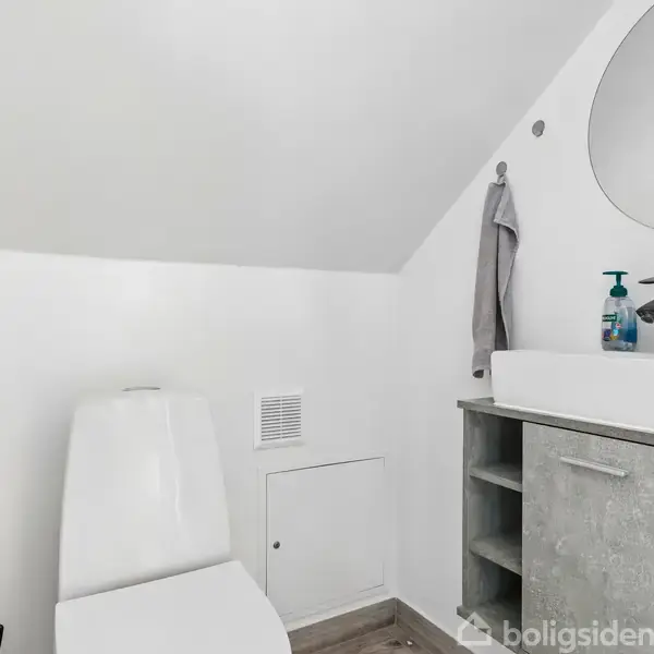 Toilet står i et lyst badeværelse under et skrånende loft med en håndvask på bænk ved siden af og et spejl ovenfor. En radiator er monteret på væggen. En flaske rengøringsmiddel står ved håndvasken.