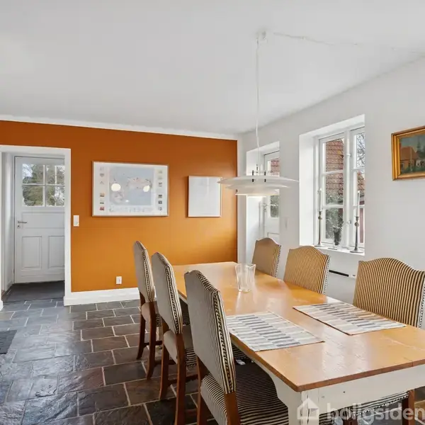 Spisebord med syv stole står i et lyst rum. En lampe hænger over bordet. Rummet har tre vinduer, en orange væg, og en dør fører til en anden del af huset. En plante er placeret i hjørnet.
