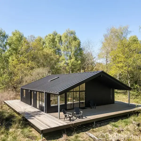Et sort sommerhus med store vinduespartier står på en træterrasse. To stole og et lille bord står på terrassen. Omgivet af tæt, grøn skov på en solrig dag.