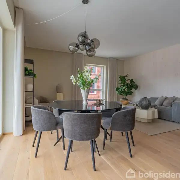 Et moderne rum indeholder et rundt spisebord med seks sorte stole, en lys buket i midten. Sofaområde i baggrunden med grønne planter. Store vinduer slipper naturligt lys ind.