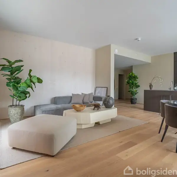 Stue med grå sofa og polstret fodskammel, placeret på et lyst trægulv. Rund spisebord med mørke stole til højre. Grønne planter som dekoration i moderne, lys lejlighed.