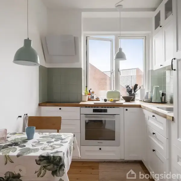 Et lyst køkken med hvide skabe og træbordplader. Et spisebord med blomsterdug står til venstre, mens en ovn og køkkenvask er ved vinduet. To hængelamper lyser rummet op.