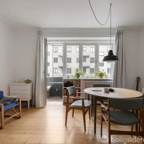 Et spisebord med stole i et lyst rum. Et vindue i baggrunden har udsigt til en bygning. Væggen har kunst, og der er planter på radiatoren.