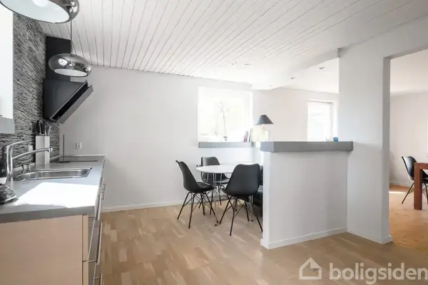 Et moderne køkken med grå skabe og en sort emhætte, står ved siden af en lille spiseplads med sorte stole. Rummet har lyse trægulve og hvide vægge.