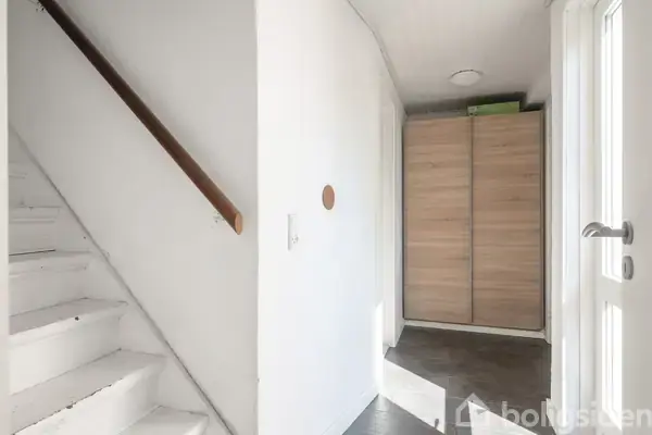 En hvid trappe med en brun håndliste fører op fra en gang med flisegulv. Til højre ses en dør, og for enden er et skab med trælook.