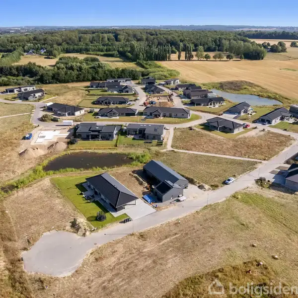 Boligområde med moderne huse set fra oven, omgivet af marker og skovområder. Husene ligger langs buede veje, og nogle grunde har små søer. Klart vejr giver en bred udsigt.