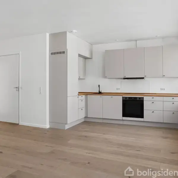 Køkken med lysegrå skabe og indbygget ovn, stående i et rummeligt, lyst rum med trægulv og store vinduer, der giver naturligt lys. Ingen tekst er til stede.