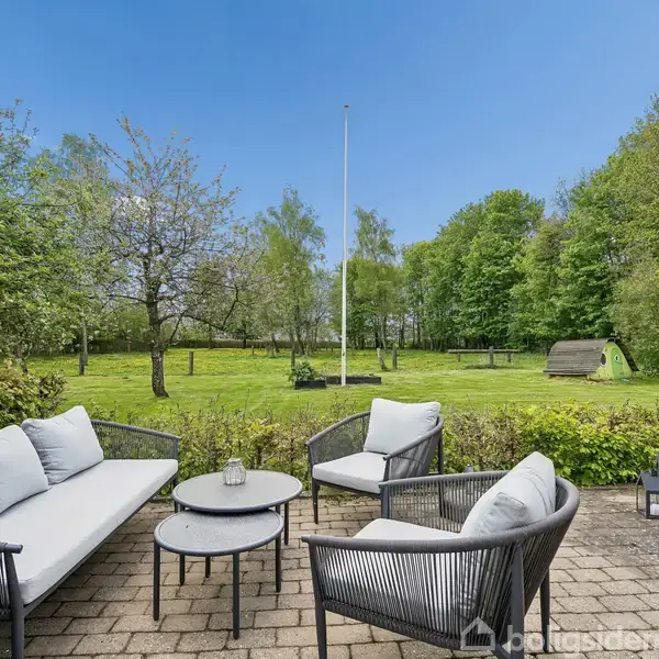 Terrassemøbler står på en stenbelagt terrasse omgivet af frodige træer og græsplæne. I baggrunden ses en flagstang og en lille legeplads. Vejret er solrigt og himlen er blå.