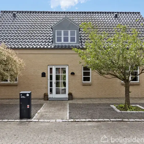 En murstensvilla med gråt tegltag står på en flisebelagt grund med en indkørsel. To træer flankerer indgangen, mens en postkasse står til venstre ved flisegangen.
