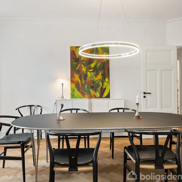 Et rundt bord med sorte stole står i et spisestueområde. En moderne, cirkulær lampe hænger over bordet. Væggen har et farverigt maleri. Rummet er lyst og minimalistisk.