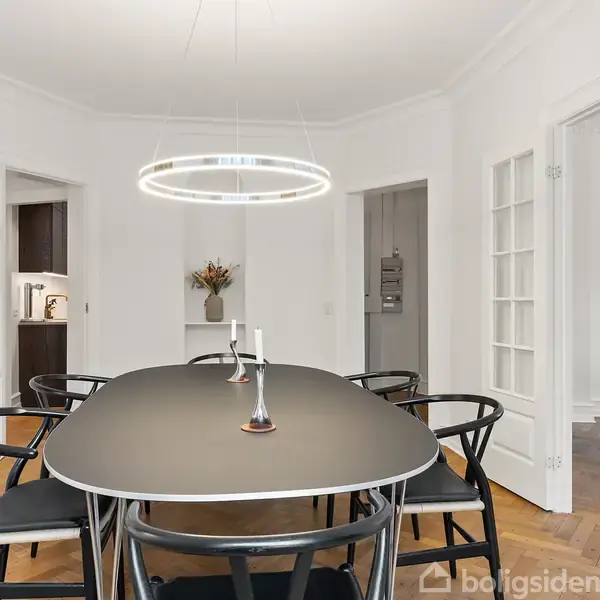 Et sort spisebord med otte stole omkring står under en moderne, rund loftslampe i et lyst spisestue. Rummet har parketgulv og hvide vægge, med åbne døre til tilstødende rum.