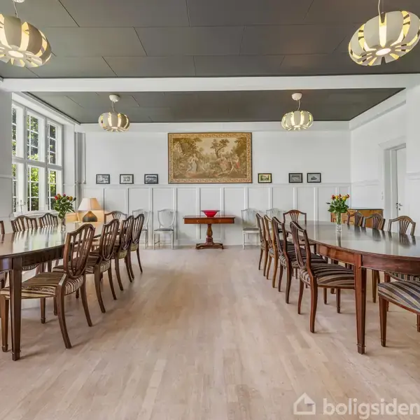 Store træborde og matchende stole er arrangeret i et lyst rum med hvide vægge og ophængte lamper. Store vinduer til venstre giver naturligt lys. Vægkunst og planter tilføjer dekoration.