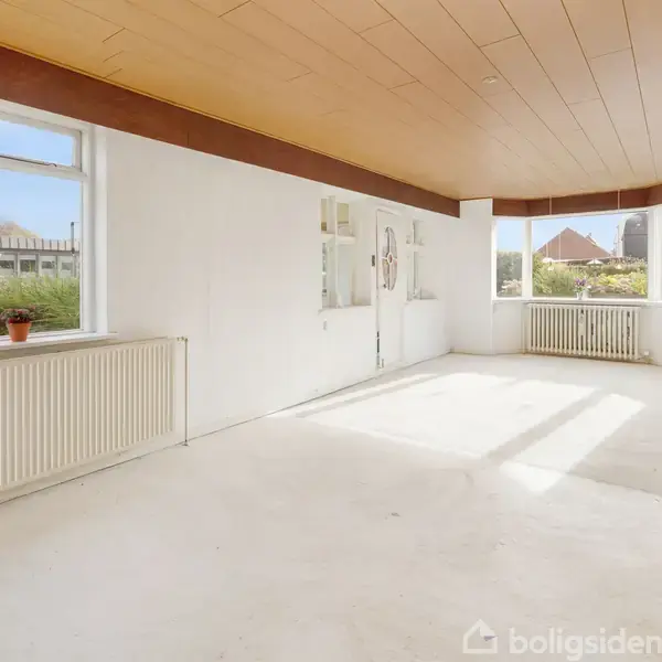 Et tomt, lyst rum med hvide vægge og gulv. Vinduet har udsigt til en grøn have. Træloftet skaber en varm kontrast. En enkelt plante står på radiatoren.