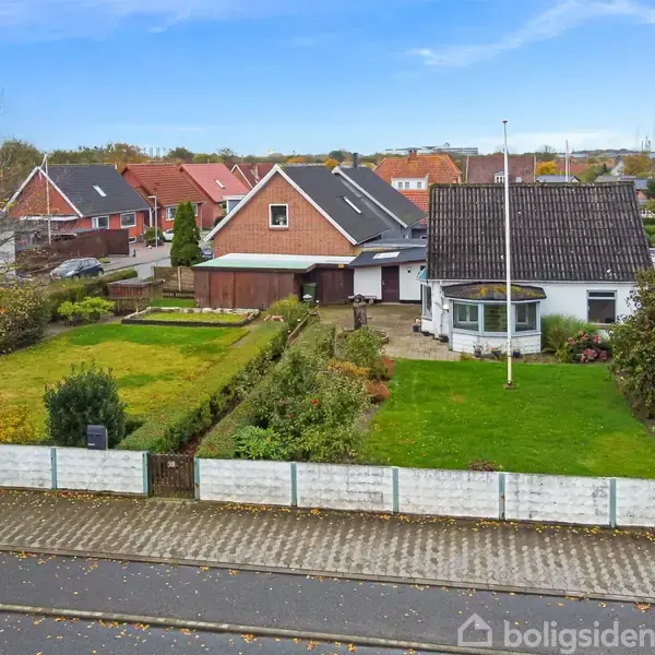 Et rødt murstenshus med en muret karnap ses fra gaden, omgivet af grønne haver og hegn. Flere villaer er synlige i nabolaget under en klar blå himmel.