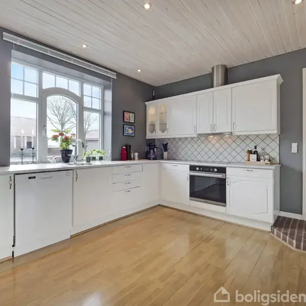 Et moderne køkken med hvide skabe og integrerede apparater. Store vinduer lader naturligt lys strømme ind. Væggene er grå, og gulvet er lyst træ. Der er ingen tekst.