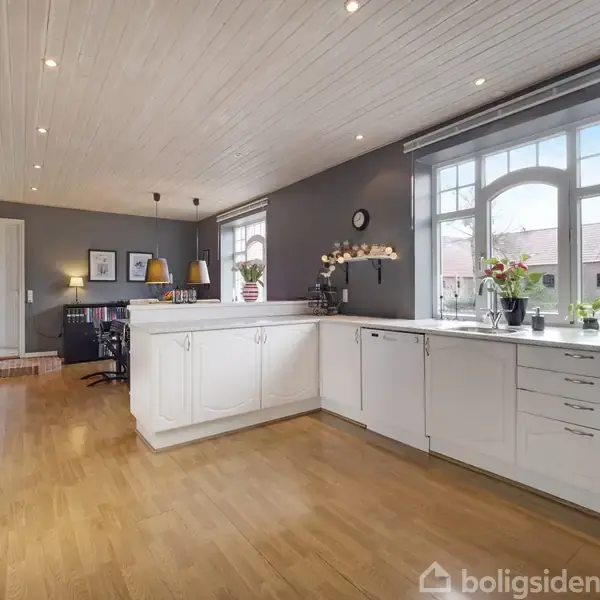 Køkken med hvide skabe og skuffer i aktivitet, dekoreret med blomster og ur, i et lyst rum med trægulv og store vinduer, der giver et naturligt lysindfald.