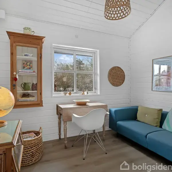 En hyggelig stue med en blå sofa og farverige puder står til højre, mens et lille bord og stol er placeret under et vindue. Rummet har også opslagstavler og et skab.