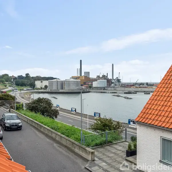En by med en havn set fra en gade. Biler står parkeret langs vejen. Orange tagsten pryder husene i forgrunden. En industriel havn og vand er i baggrunden.