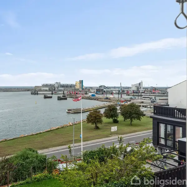 Udsigt over en havn med kajer og både, set fra et vindue. Til venstre et hus med rødt tag, og i baggrunden himmel og vand.