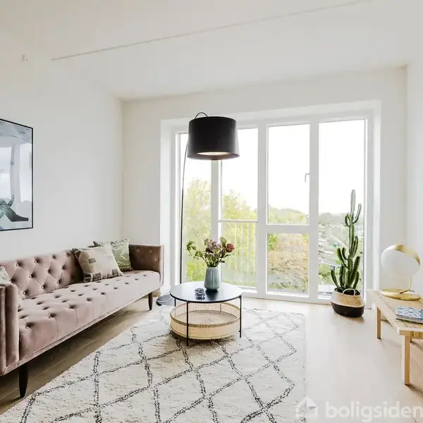 Brun sofa står i stuen, flankeret af et rundt sofabord med blomster på, foran store vinduer. Et tæppe ligger på gulvet, og en kaktus står til højre.