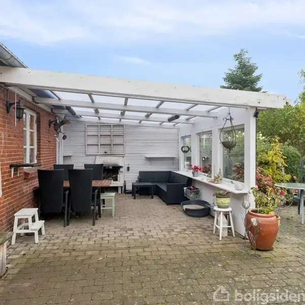 Overdækket terrasse i en have. Møbler med sort spisebord og stole under hvid træpergola. Røde murstensvægge til venstre, potteplante og havemøbler til højre. Grønt buskads omkranser området.