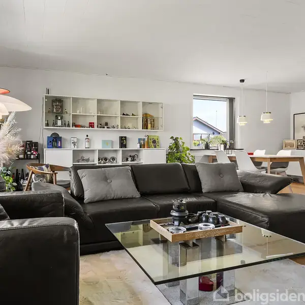 Sort lædersofa står stille i en moderne, lys stue med store vinduer. Reol og spisebord med stole er synlige i baggrunden, omgivet af indrammet kunst og dekorative genstande.