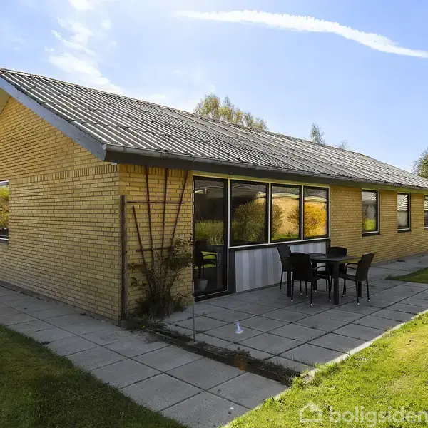 Et gult murstenshus står stille på en græsplæne, omgivet af fliser; foran huset står et bord med fire stole.