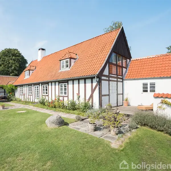 Et bindingsværkshus med rødt tegltag står i en velplejet have med grøn græsplæne og buske. Solen skinner, og der er en lille terrasse foran huset.