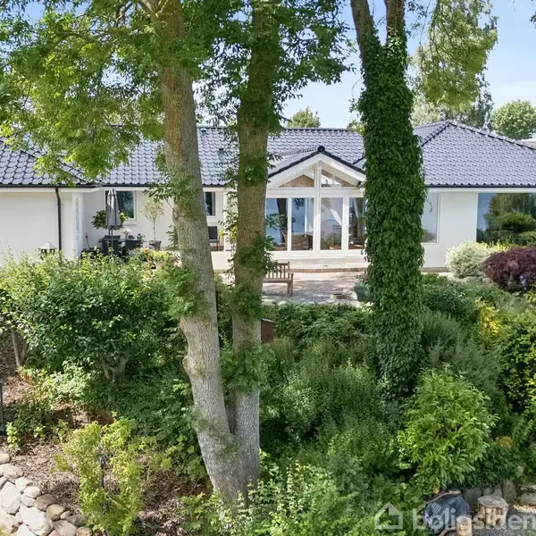 Huset er hvidt med et sort tegltag, omgivet af frodig grøn vegetation og en velplejet have. Flere store træer står foran huset, der har store vinduer og terrasse.