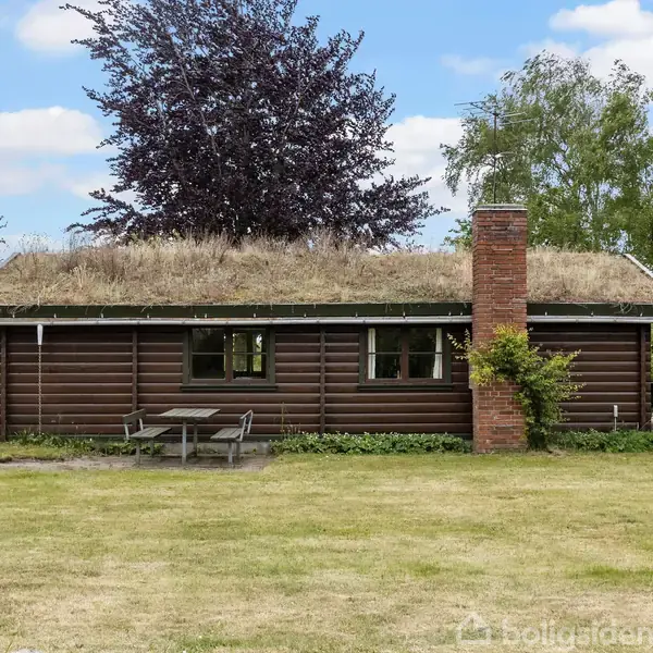 En træhytte med græs på taget står stille i en have med grønne træer og buske omkring. En bordbænkesæt er placeret foran hytten.