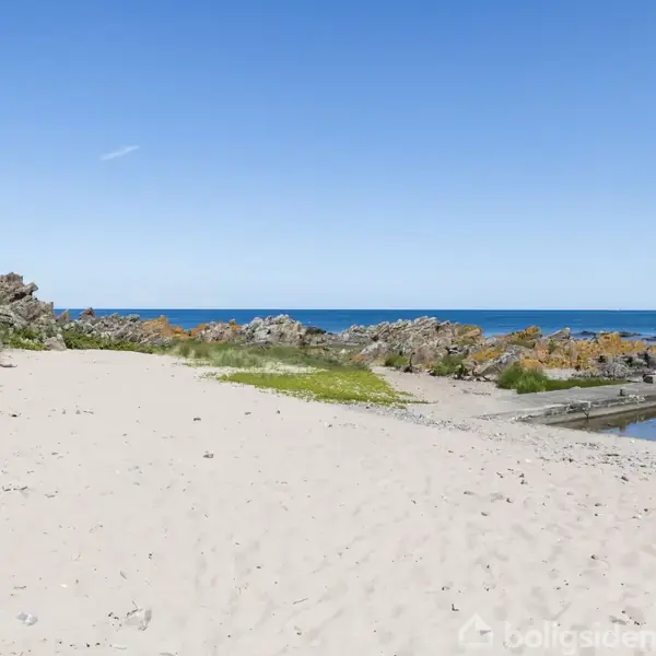 Sandstrand strækker sig mod klipper med græs ved kanten, under en klar blå himmel. Havet skimtes i horisonten. Ingen tekst fundet i billedet.