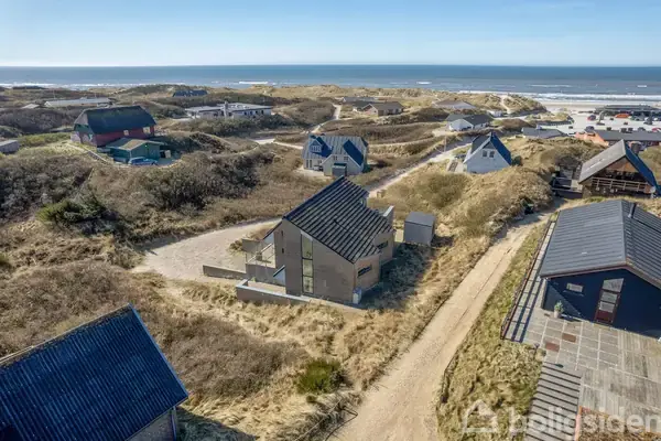 Et moderne hus står stille midt i klitterne nær kysten. Flere huse er spredt omkring i det sandede, grønne landskab, med havet svagt glitrende i baggrunden under en klar himmel.