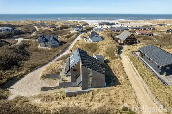 Et moderne hus med store vinduespartier står blandt klitterne ved kysten. Huset er omgivet af sandstier og andre spredte boliger med stranden og havet i baggrunden.