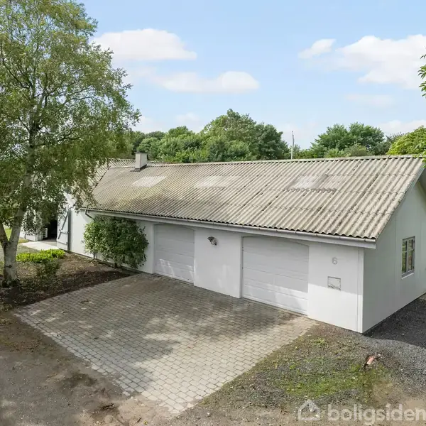 En hvid garagebygning med to porte står stille omgivet af træer og en grussti i et grønt, landligt område.