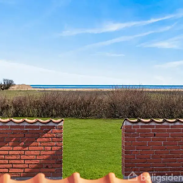 Mur af røde mursten står stille, omkranset af en grøn græsplæne, med udsigt til en kystlinje og et blåt hav i baggrunden under en klar himmel.