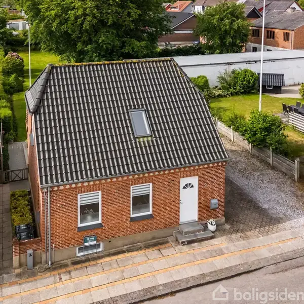 En murstenshus med sort tegltag og kvistvindue står stille i en velplejet forstadshave med græsplæner og buske. Der er en hvid dør og to vinduer foran.