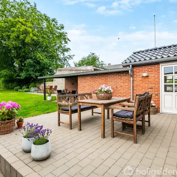 Træmøbler står på en brolagt terrasse foran et murstenshus med en hvid dør. Farverige blomster i potter står spredt omkring. Græsplæne og træer ses i baggrunden.