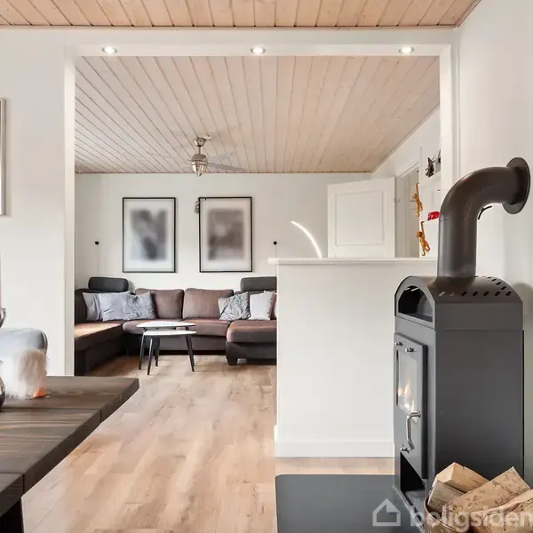 En moderne brændeovn står i en lys stue med trægulv og loft. Sofa og billeder ses i baggrunden. Et bord med lysestager står i forgrunden. Plakat på væggen.