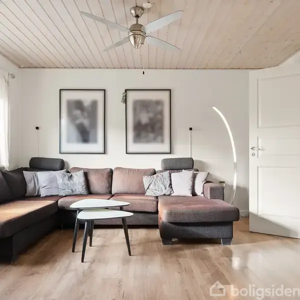 En stor, brun hjørnesofa står i en lys stue med træloft og trægulv. To billeder hænger på væggen, og en loftsventilator er synlig. En åben dør fører til et andet rum.