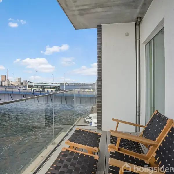 To træstole står på en balkon med glassider og udsigt over vandet mod en by i det fjerne, under en skyfri blå himmel.
