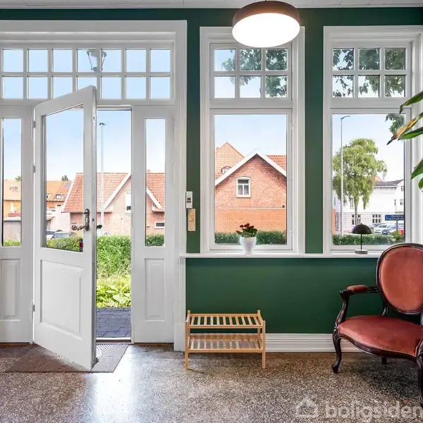 Entré med åbent dør. Grønne vægge, gyngestol og plante ved vinduet. Udenfor ses huse og træer. Gulvet er terrazzo, og der er en skohylde ved væggen.