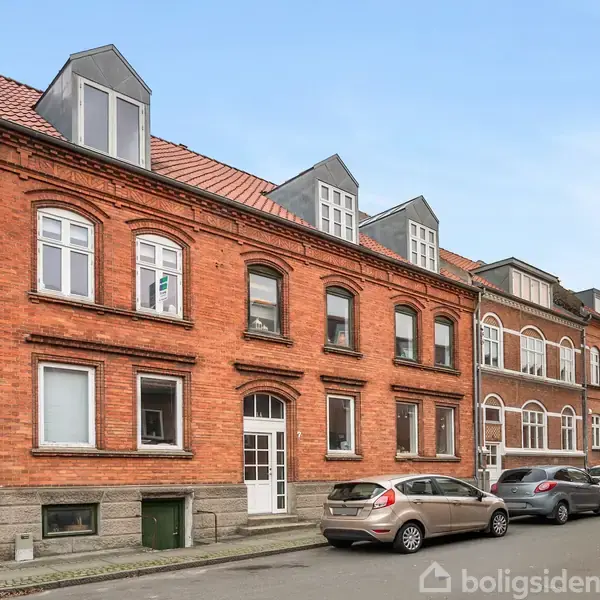 Røde murstensbygninger med vindueskarnapper står langs en stille gade. Flere parkerede biler holder på vejen. Klar himmel over bygningerne uden tydelig menneskelig aktivitet.