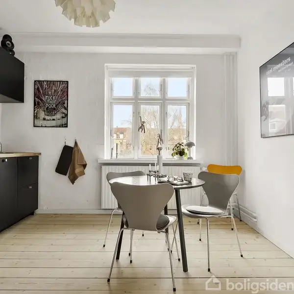 Et køkken med sorte skabe langs venstre væg, en rund bord med to stole foran et vindue. Lyse trægulve og en minimalistisk indretning med kunst på væggene.
